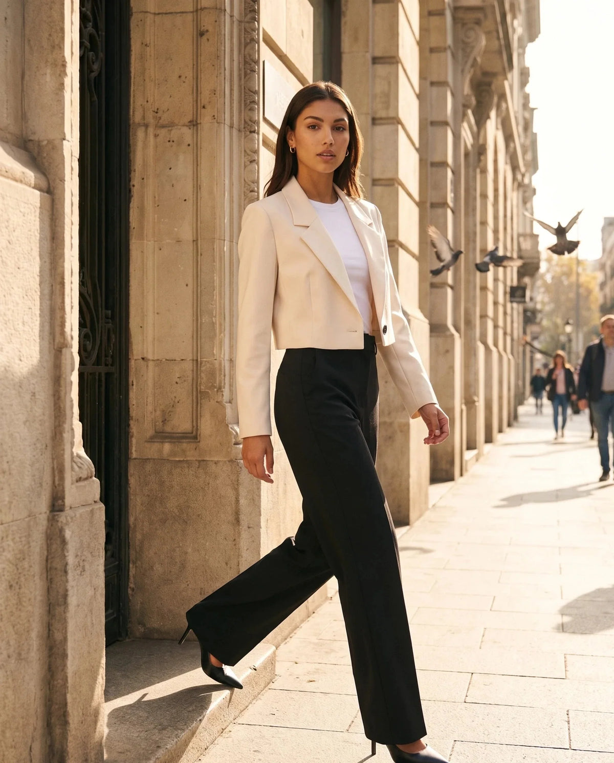 Blazer court beige Léontine, porté devant un immeuble en pierre calcaire au soleil