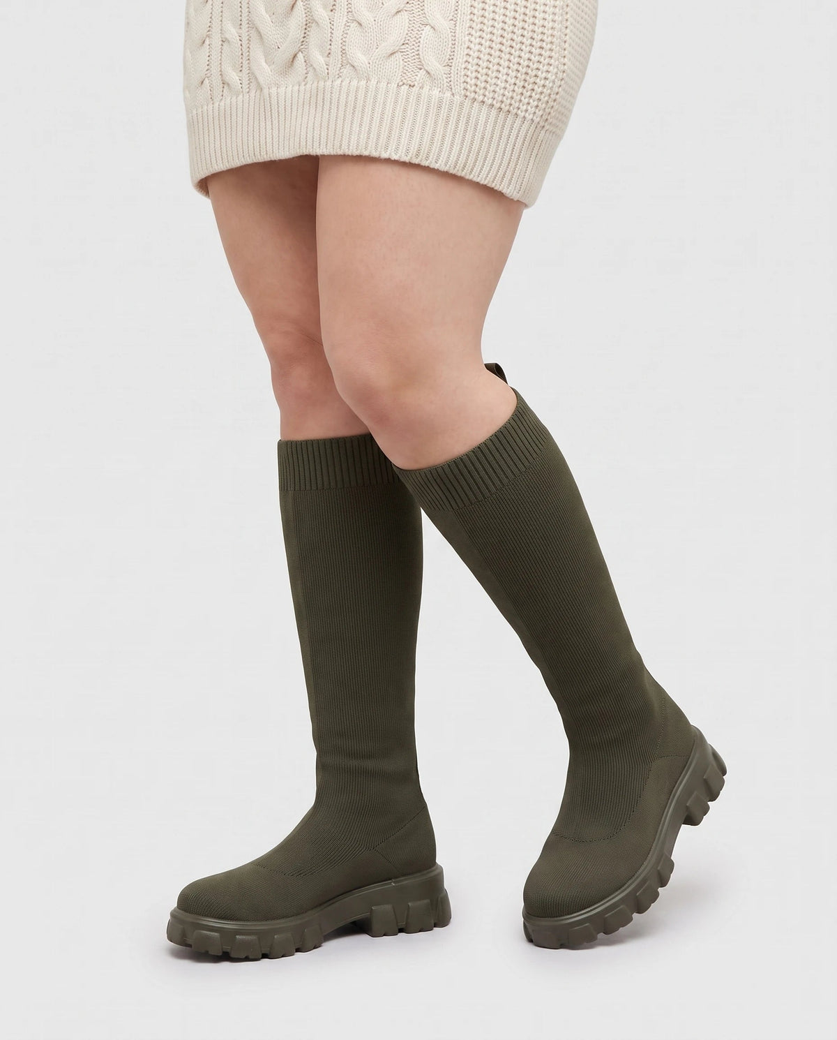 Bottes chaussettes hautes en maille Sylvette — Kaki, portées en plan rapproché avec une robe pull écrue
