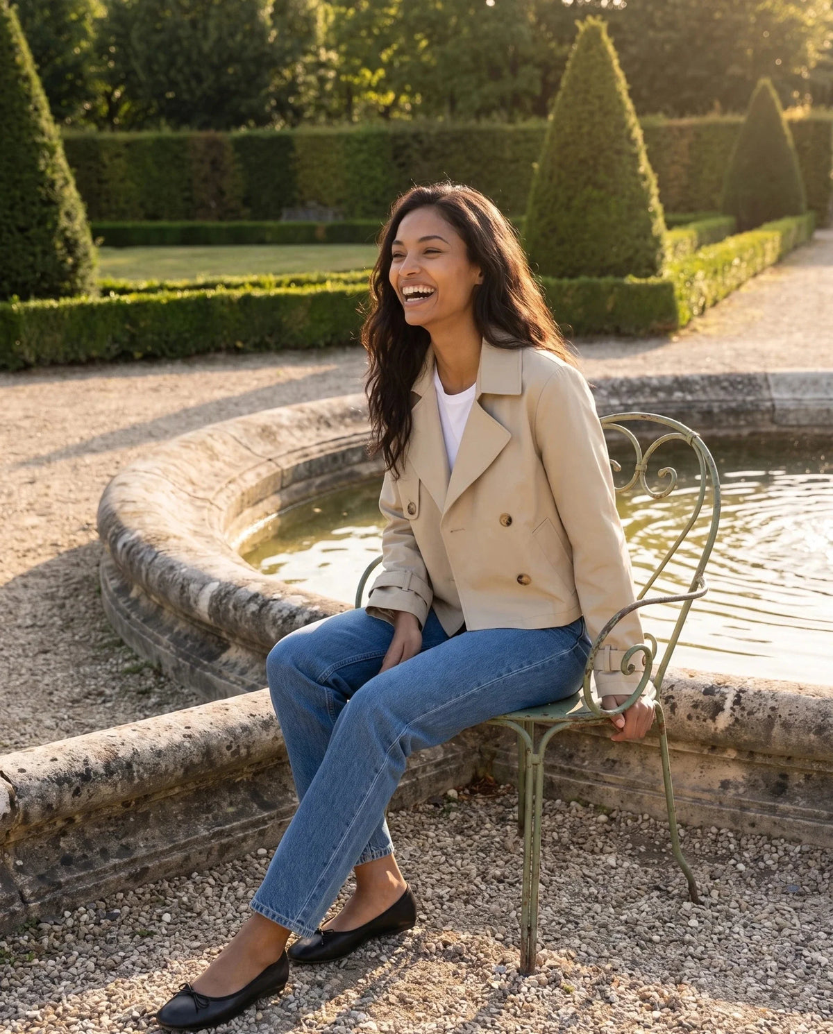 Trench court croisé beige Aliénor, porté près d'une fontaine de jardin à la française