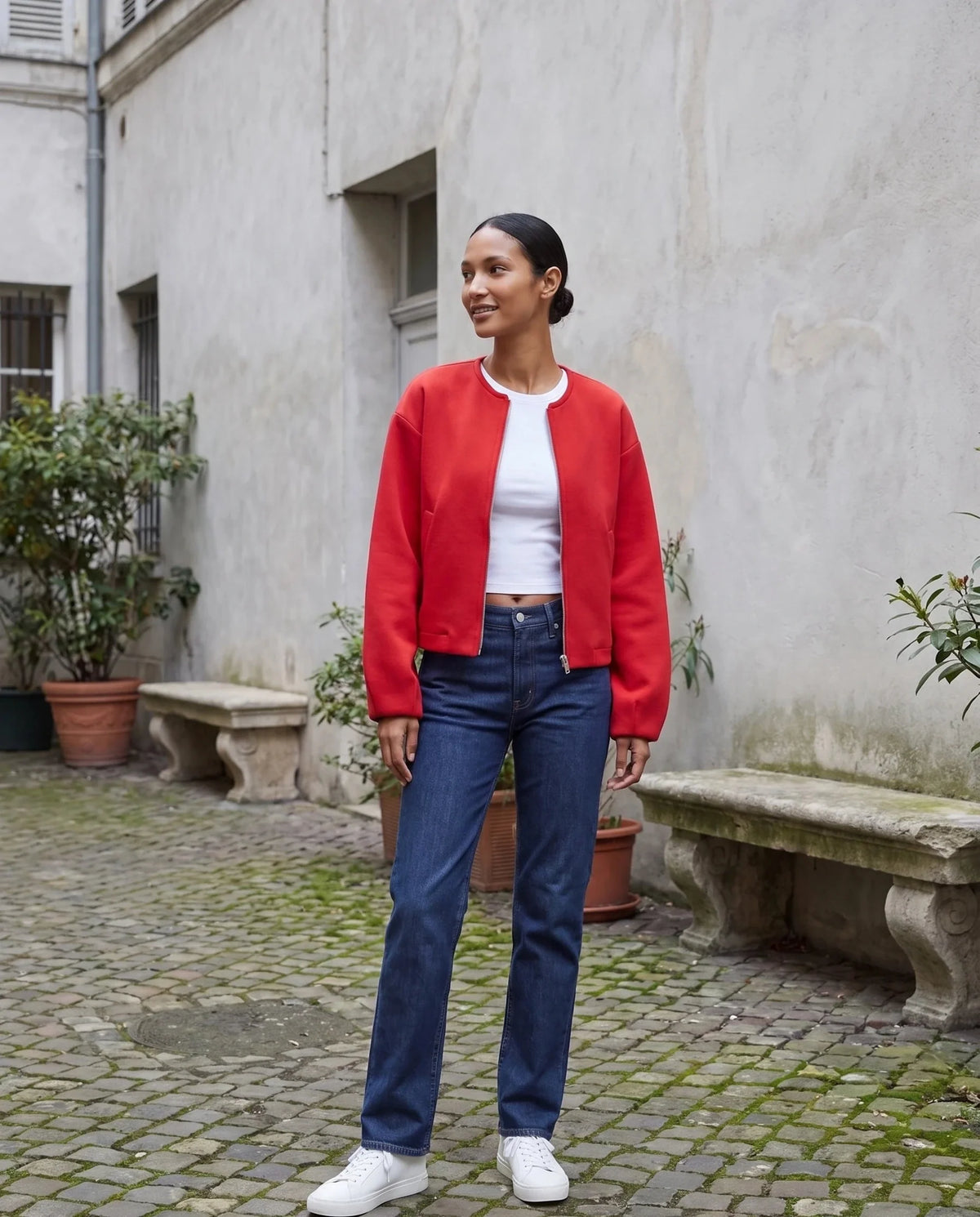 Veste zippée sans col rouge Capucine, portée dans une cour aux murs gris perle